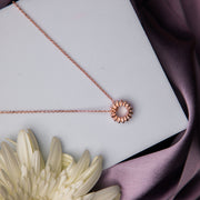 A rose gold-finish pendant chain featuring a sundial design, displayed on a white surface with a purple fabric backdrop and a flower.